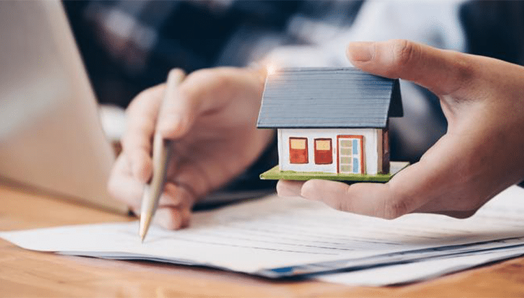 person writing on a paper while holding a mini house toy