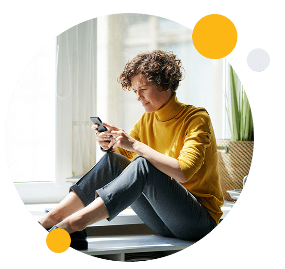 Calm curly-haired woman in sweater sitting on shelf near paper container and using smartphone