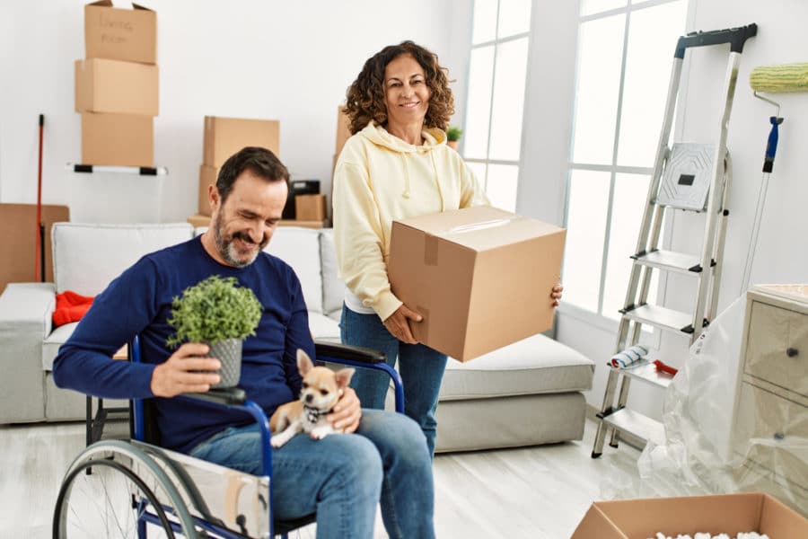 Middle age hispanic couple smiling happy. Man sitting on wheelchair with dog on his legs and woman holding cardboard box at new home. stock phot