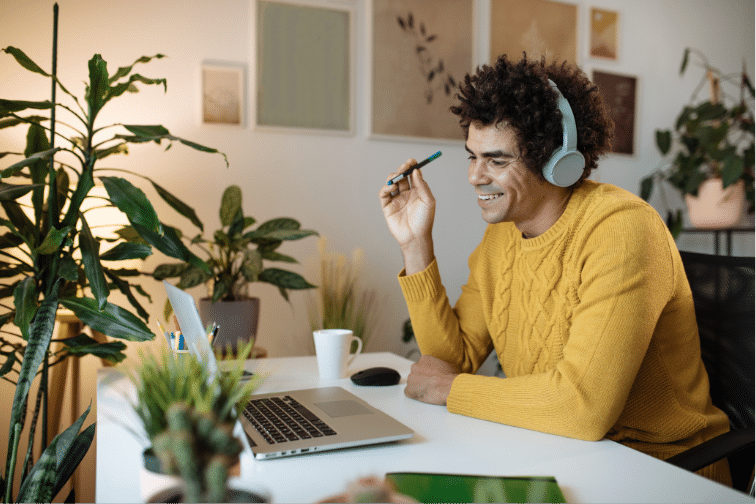 A person in a yellow sweater and headphones sitting at a desk with a laptop, a cup, and various plants around in a room with art on the walls.