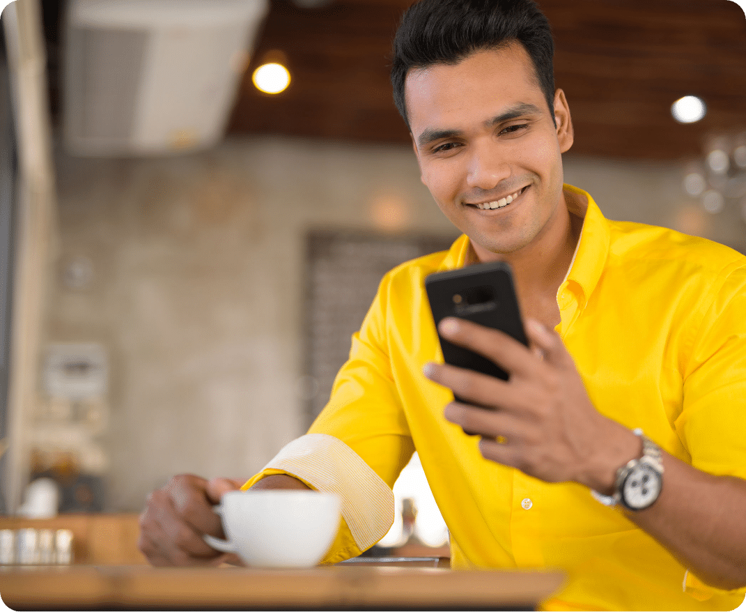 Young happy Indian man using phone and sitting by the window at café