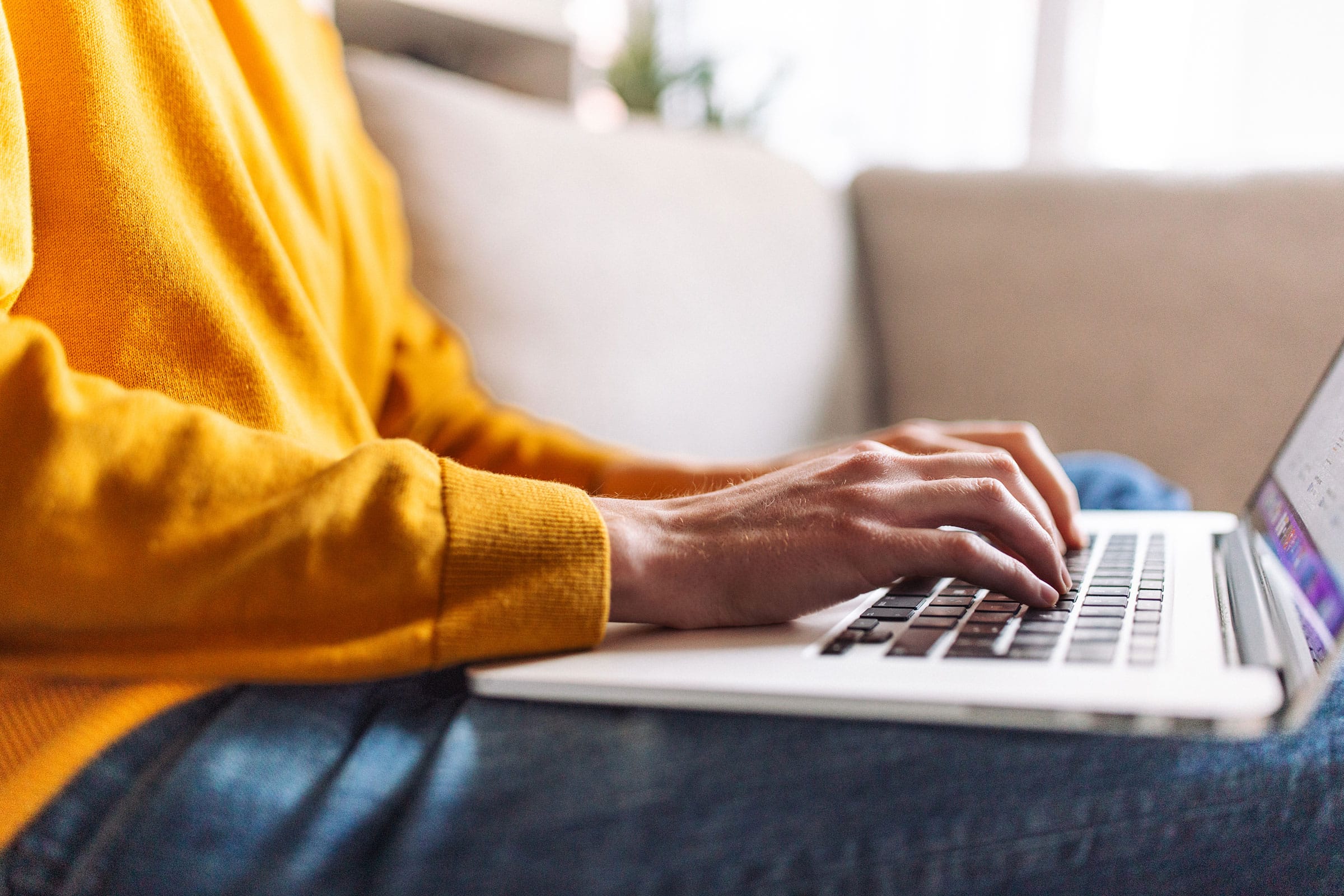 A woman wearing yellow shirt having an enquiry using a laptop, sitting comfortably on a couch in a homely environment with warm lighting